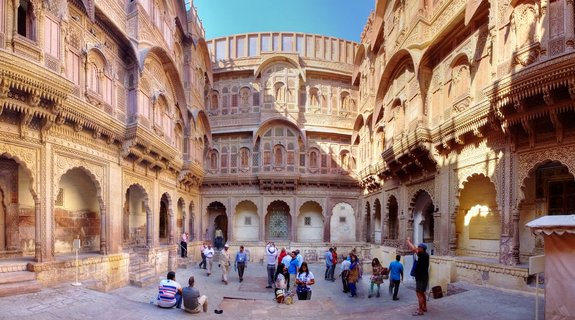 Wide courtyard surrounded by multi-story golden sandstone building with ornate carved balconies, arched walkways, and tourists walking on stone pavement under clear blue sky