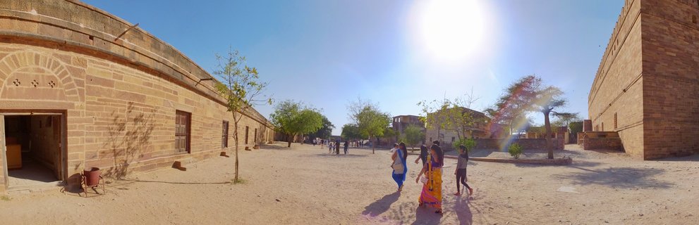 Wide courtyard between yellow sandstone buildings in Jaisalmer, India with people walking including women in colorful saris under bright sun and clear blue sky