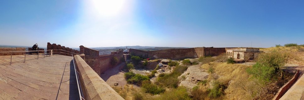 Panoramic view of Jaisalmer Fort showing golden sandstone walls, courtyard, metal railings along walkway, desert vegetation, and clear blue sky with bright sun