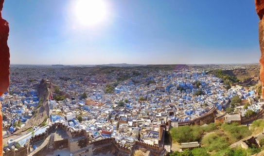 Panoramic aerial view of densely packed blue and white buildings of Jodhpur's old city with fort walls in foreground and arid hills in the distance under bright blue sky