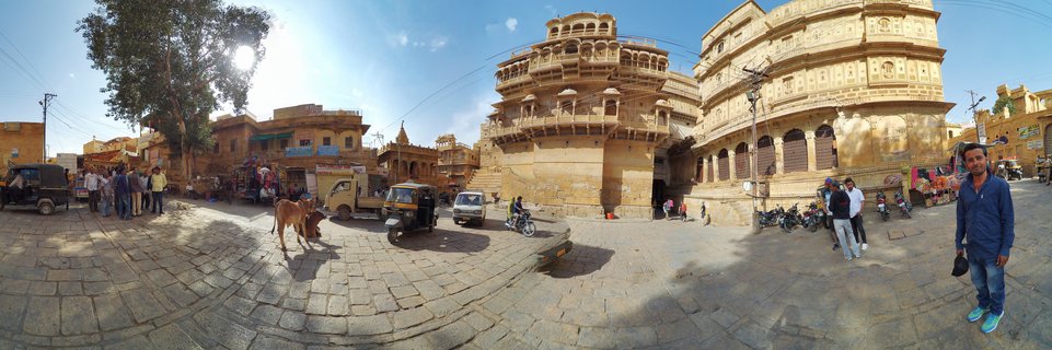 Panoramic street view of Jaisalmer showing yellow sandstone buildings with ornate balconies, people walking, auto-rickshaws, motorcycles, and a cow on cobblestone pavement under blue sky