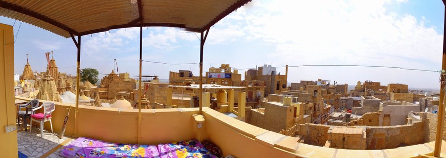 Panoramic view from a covered rooftop terrace overlooking golden sandstone buildings and temple spires in Jaisalmer, India, with blue sky and white clouds above