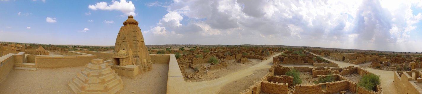 Ancient Hindu temple with golden sandstone tower surrounded by desert ruins and sparse vegetation under cloudy blue sky in Jaisalmer, India