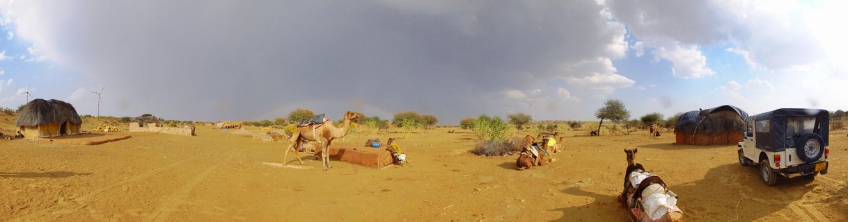 Panoramic desert scene showing multiple camels on sandy ground with traditional thatched huts, a white utility vehicle, wind turbines in the distance, and dramatic storm clouds with rainbow over Jaisalmer, India