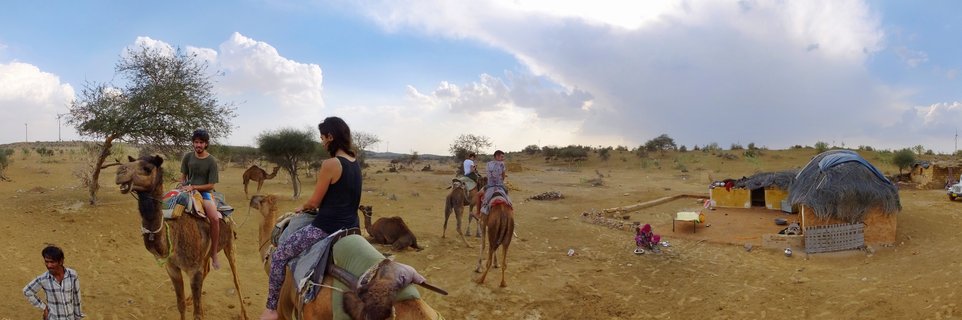 Panoramic view of tourists riding camels in the Thar Desert with traditional huts and acacia trees under a blue sky with clouds