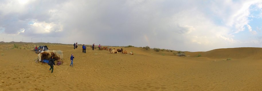 Panoramic view of sand dunes with tourists, camels, and desert camp tents under cloudy sky in Thar Desert, India