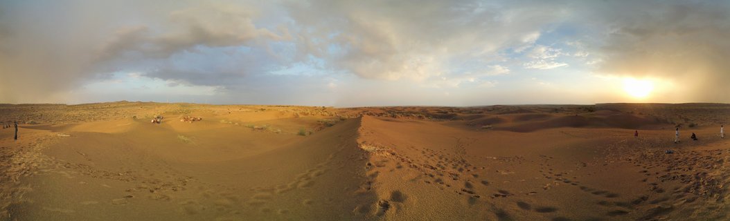 Panoramic desert landscape showing golden sand dunes under a cloudy sky with the sun setting on the horizon, with small human figures and footprints visible across the sandy terrain