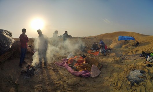 A desert camp scene in Jaisalmer with people gathered around campfires on sand dunes, colorful blankets spread on the ground, and temporary tarp shelters under a clear morning sky