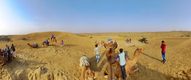 Wide panoramic view of tourists riding camels across golden sand dunes in the Thar Desert near Jaisalmer, India, with clear blue sky and desert vegetation visible