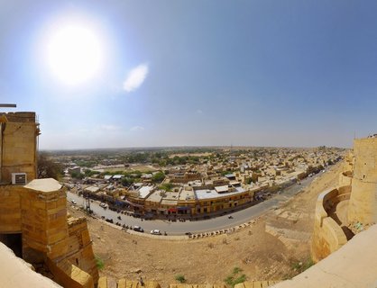Wide panoramic photograph showing the golden-colored city of Jaisalmer, Rajasthan, India, viewed from an elevated fort position. Yellow sandstone buildings fill the middle ground with a curved road and vehicles visible below. The bright sun appears in the upper left of a clear blue sky, with desert terrain extending to the horizon.