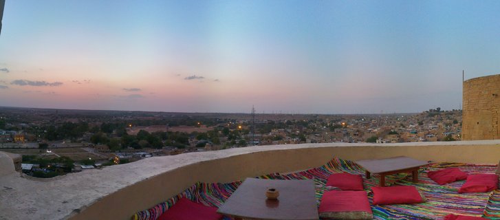 Panoramic view from a rooftop terrace with colorful rugs, wooden tables and red cushions overlooking Jaisalmer city at dusk with golden stone buildings and pink-blue sky