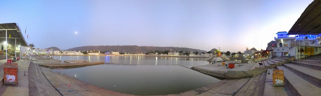 Panoramic view of Pushkar Lake at dusk showing stone steps leading to water, illuminated buildings, distant hills, and moon in blue sky