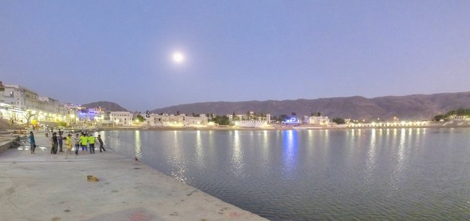 Panoramic view of Pushkar Lake at twilight with pilgrims standing on stone ghats, illuminated buildings along the shoreline, moon in the purple-blue sky, and hills in the background with reflections visible in the water.