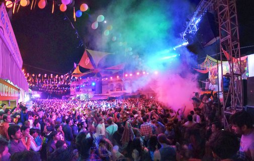 Night scene of Holi festival celebration in Pushkar, India showing massive crowd throwing colored pink, blue and green powder into the air. Professional stage with lighting truss visible on right, balloon strings overhead, and illuminated food vendor stalls on left side.