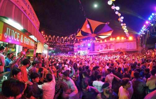 Large crowd of people dancing at night festival in Pushkar India with colorful purple and blue stage lights, decorative lanterns overhead, and food stalls visible