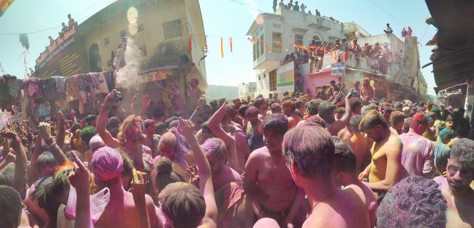 Large crowd of people covered in pink and purple colored powder celebrating Holi festival on a street in Pushkar, India with buildings and spectators on rooftops