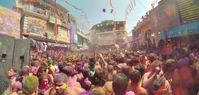 Large crowd of people covered in bright colored powders during Holi festival celebration in a street square with buildings and banners in Pushkar, India