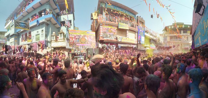Large crowd of people covered in colored powder celebrating Holi festival in Pushkar street with buildings and banners overhead
