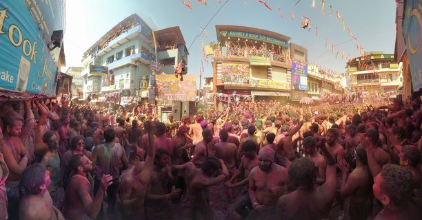 Large crowd of people covered in pink and purple colored powder celebrating Holi festival in street of Pushkar India with buildings and colorful banners visible