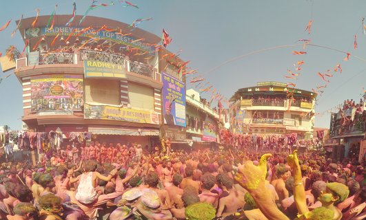 Large crowd of people covered in bright colored powders celebrating Holi festival in street of Pushkar, India with buildings and banners in background