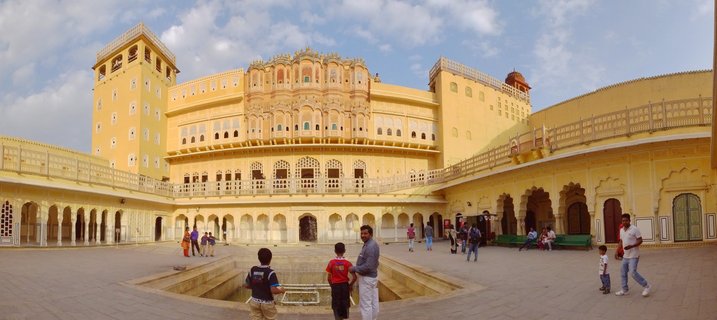 Wide view of City Palace courtyard in Jaipur showing yellow sandstone palace buildings with ornate windows, arched walkways, and tourists walking on stone pavement