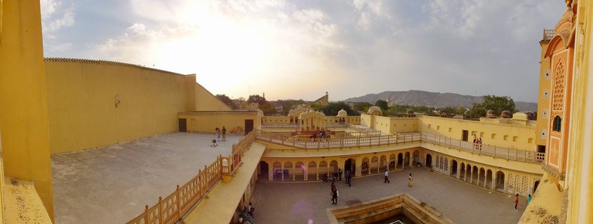 Aerial view of Amber Fort courtyard in Jaipur showing yellow sandstone architecture with arched colonnades, tourists walking on multiple levels, and mountains visible in the background under partly cloudy sky