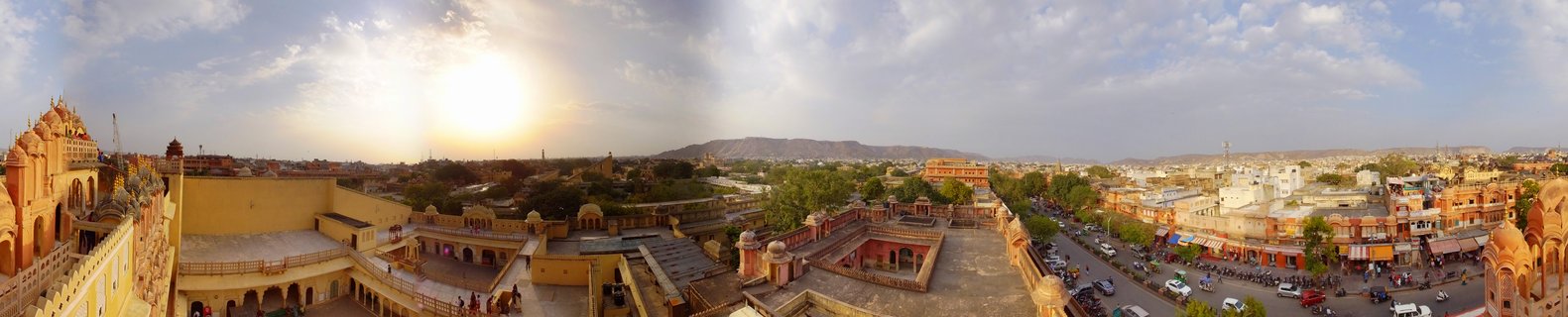Panoramic view from Hawa Mahal palace rooftop showing ornate Rajasthani architecture, busy city streets with vehicles and pedestrians, and distant hills under a partly cloudy sky at sunset in Jaipur, India