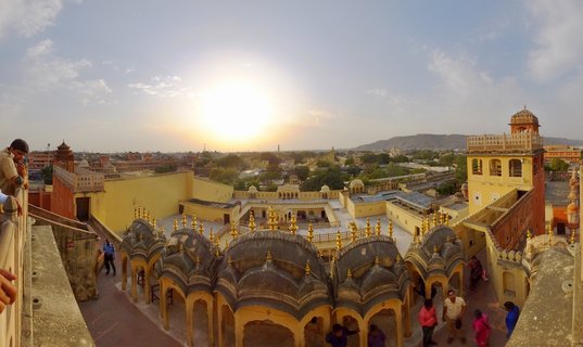 Aerial panoramic view of Hawa Mahal palace complex in Jaipur showing ornate domed structures with golden spires, yellow sandstone courtyards, tourists walking on terraces, and the sun setting over distant hills