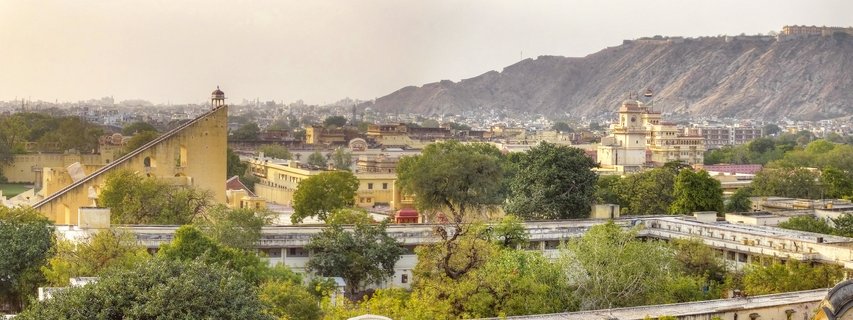 Panoramic cityscape showing the triangular Samrat Yantra sundial of Jantar Mantar observatory in foreground, traditional Rajasthani buildings, green trees, and Nahargarh Fort on the Aravalli Hills in the background under hazy sky