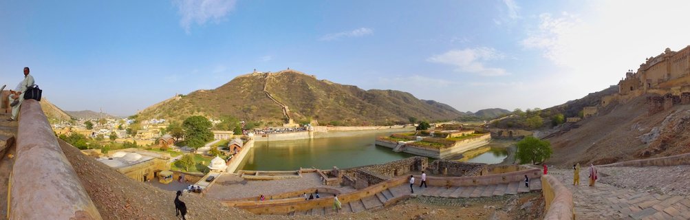 Wide panoramic photograph showing Amber Fort palace complex with Maota Lake in center, fortified walls climbing hills, tourists on stone walkways, clear blue sky above Rajasthan landscape