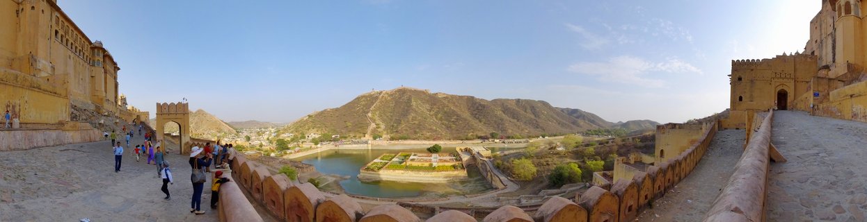 Panoramic view of Amber Fort in Jaipur showing yellow sandstone fortress walls with tourists walking on ramparts, Maota Lake with garden island below, and Aravalli Hills in the background under blue sky
