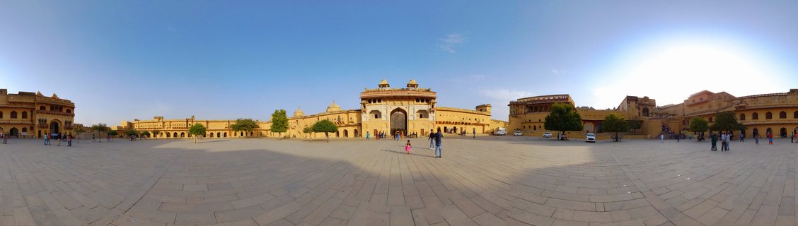 Wide panoramic photograph of Amber Fort showing golden sandstone palace buildings surrounding a large paved courtyard with tourists walking, under a clear blue sky in Jaipur, India