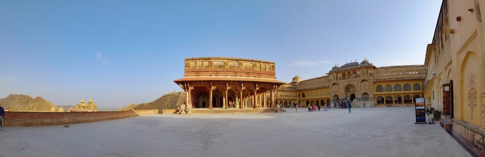 Wide courtyard at Amber Fort showing the Diwan-i-Aam pavilion with red sandstone columns, palace buildings, and visitors walking on the stone courtyard under blue sky
