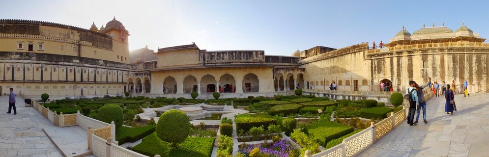 Wide courtyard of Amber Fort showing Mughal-style formal gardens with geometric green hedges, surrounded by sandstone palace buildings with domes and arches, visitors walking on stone pathways under clear blue sky