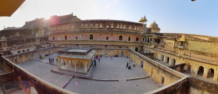Panoramic view of Amber Fort courtyard showing yellow sandstone palace buildings with multiple levels, arches, domed pavilions, and tourists walking on stone courtyards under clear blue sky with sun flare