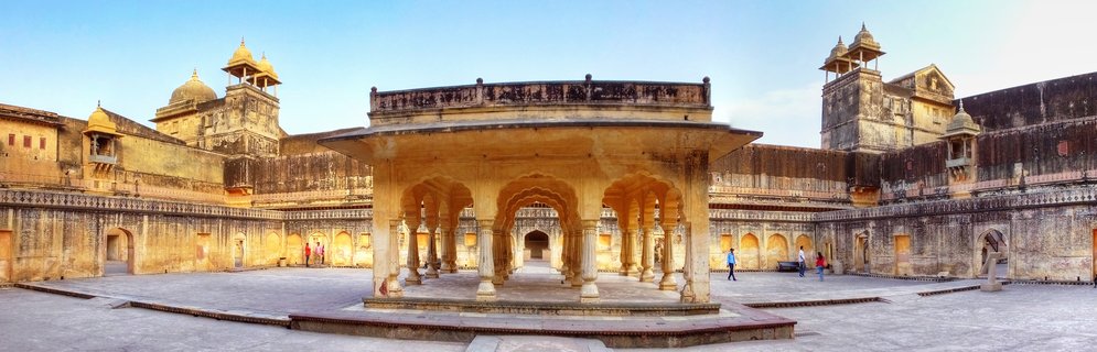 Historic sandstone courtyard with central pavilion featuring multiple arched columns at Amber Fort in Jaipur, India, with visitors walking on the stone plaza surrounded by palace walls and domed structures