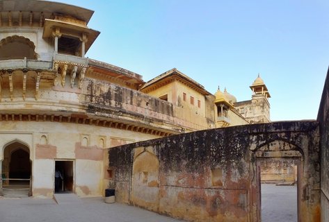 Historic Indian palace courtyard with weathered yellow sandstone walls, ornate balconies with carved brackets, arched doorways, and domed pavilions against clear blue sky