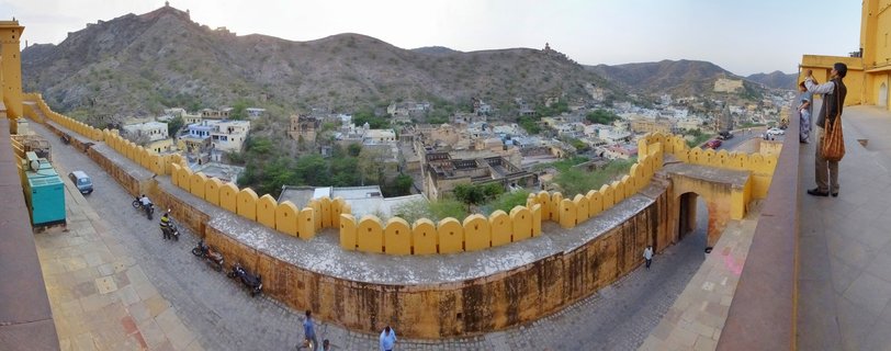 Panoramic view from Amber Fort showing yellow sandstone battlements with crenellated walls, tourists on stone walkway, town buildings below, and arid hills with fortifications in the background under soft afternoon sky