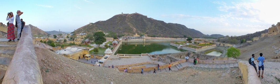 Wide panoramic photograph showing Amber Fort palace complex overlooking Maota Lake in Jaipur, India, with tourists on stone terraces and arid hills in the background