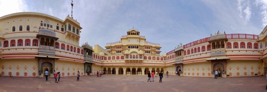 Panoramic view of City Palace courtyard in Jaipur, India, showing cream-yellow sandstone buildings with red-pink decorative elements, ornate jharokha balconies, arched Mughal windows, and tourists exploring the historic palace grounds under a clear blue sky.