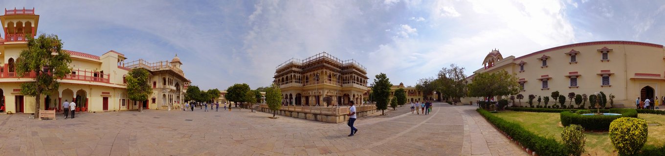 Wide courtyard surrounded by cream-colored historic buildings with red trim, arched windows, balconies, and visitors walking on stone pavement under blue sky in Agra
