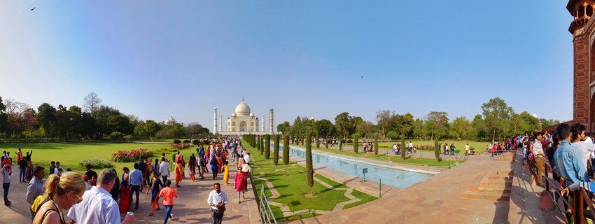 White marble Taj Mahal mausoleum with central dome and minarets viewed across a long reflecting pool, surrounded by green gardens, cypress trees, and numerous visitors on red stone pathways under clear blue sky