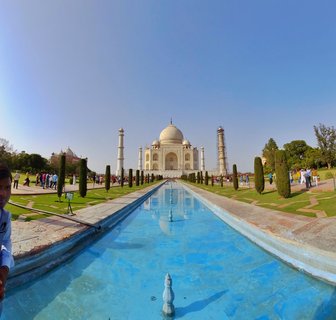 White marble Taj Mahal mausoleum with large central dome and four minarets, reflected in a long blue water channel, surrounded by green Mughal gardens with visitors walking on pathways under clear blue sky