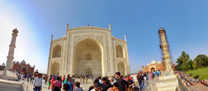 White marble gateway building of the Taj Mahal with ornate arches and Islamic patterns, flanked by two minarets, with crowds of tourists in the foreground under blue sky