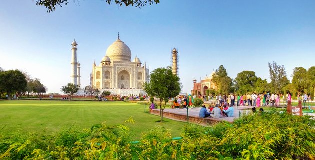 White marble Taj Mahal mausoleum with central dome and minarets surrounded by green lawns, trees, and numerous visitors under clear blue sky in Agra, India