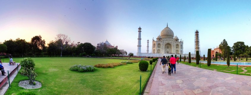 White marble Taj Mahal mausoleum with large central dome and four minarets viewed from formal Mughal gardens with green lawns, trees, and visitors walking on stone pathway during sunset