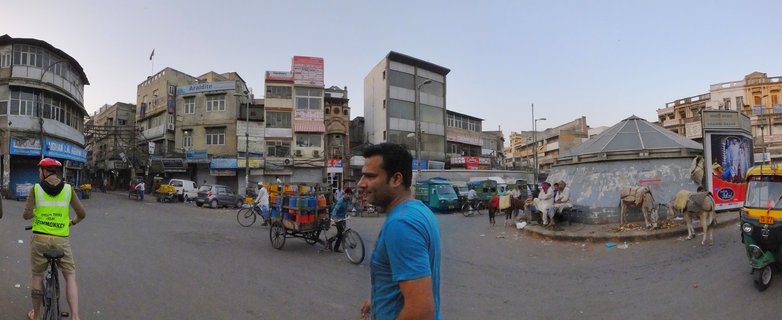 Wide panoramic street photograph showing a busy Delhi intersection with a cyclist wearing a yellow safety vest on the left, a man in blue shirt in the foreground, cycle rickshaws with colorful containers, green and yellow auto-rickshaws, multi-story buildings with signs, and a small temple structure with cows on the right side.
