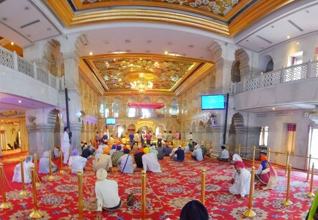 Interior view of Sikh Gurdwara prayer hall in Delhi with devotees in colorful turbans sitting on red carpet beneath ornate golden ceiling and chandelier