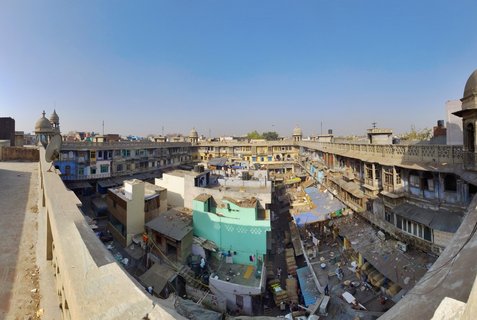 Aerial view of Old Delhi market courtyard surrounded by historic multi-story buildings with domed towers, colorful weathered facades, and people working below under clear blue sky