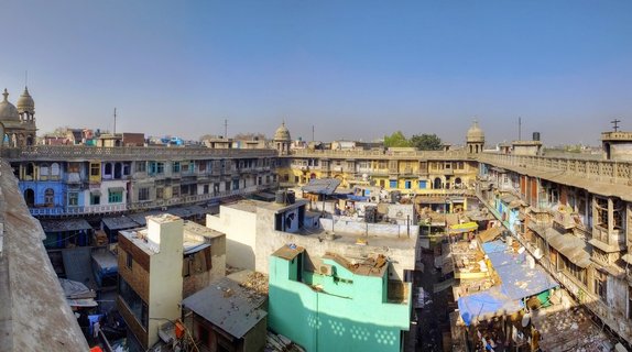 Aerial panoramic view of a historic courtyard in Old Delhi surrounded by multi-story buildings with domed towers, colorful facades in blue yellow and green, and rooftop structures under clear blue sky
